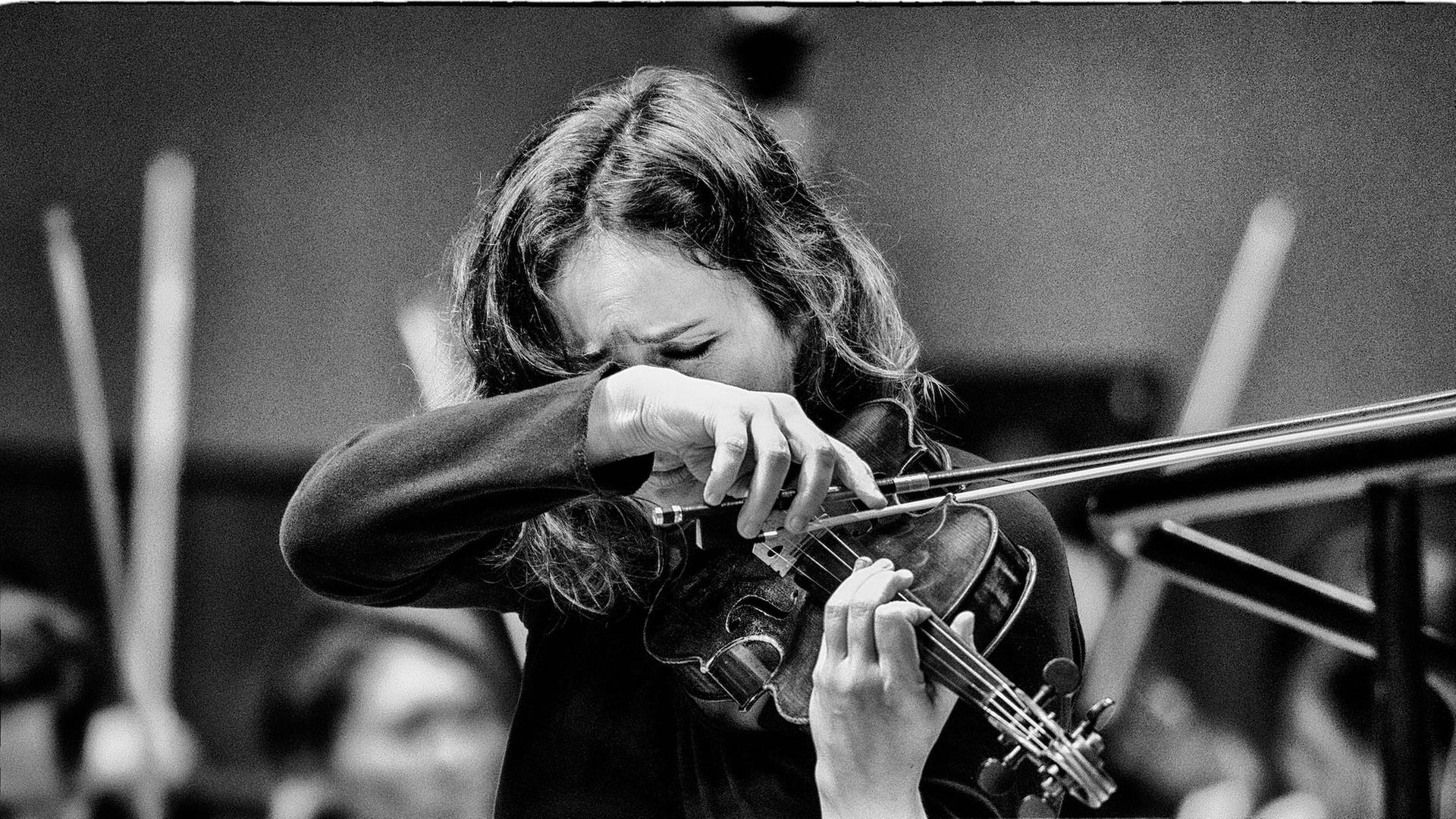 Patrica Kopatchinskaja steht hinter einem Notenpult vor einem Orchester und spielt so wild, dass ihre Haare und ihre Hände das Gesicht beinahe verdecken. 