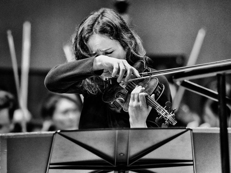 Patrica Kopatchinskaja steht hinter einem Notenpult vor einem Orchester und spielt so wild, dass ihre Haare und ihre Hände das Gesicht beinahe verdecken. 