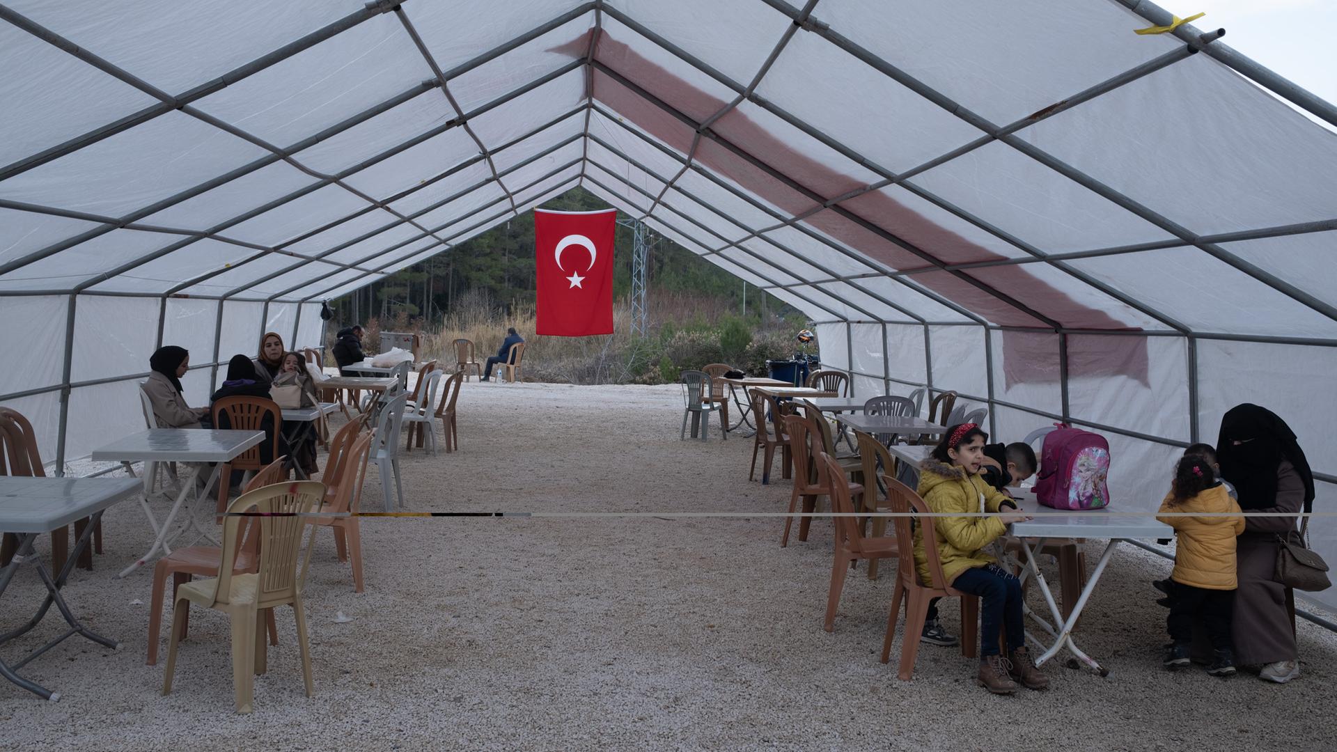 Geflüchtete sitzen in einem Zelt in dessen hinterem Teil eine türkische Flagge hängt.