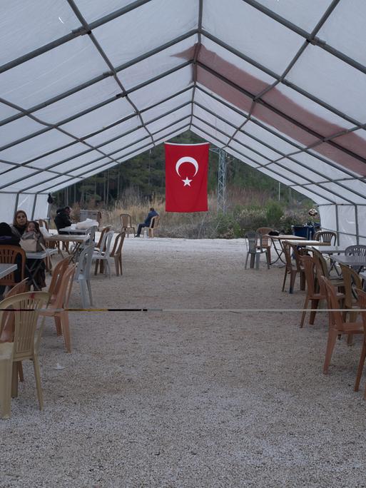 Geflüchtete sitzen in einem Zelt in dessen hinterem Teil eine türkische Flagge hängt.