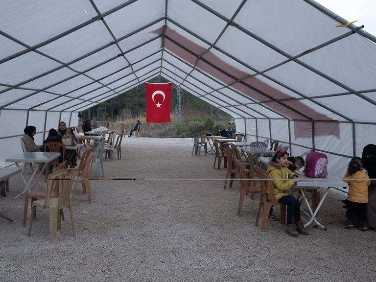 Geflüchtete sitzen in einem Zelt in dessen hinterem Teil eine türkische Flagge hängt.