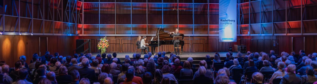 Blick in den Kammermusiksaal im Funkhaus Köln mit einer blau-rot beleuchteten Bühne, auf der ein Flötist von einer Pianistin begleitet wird.