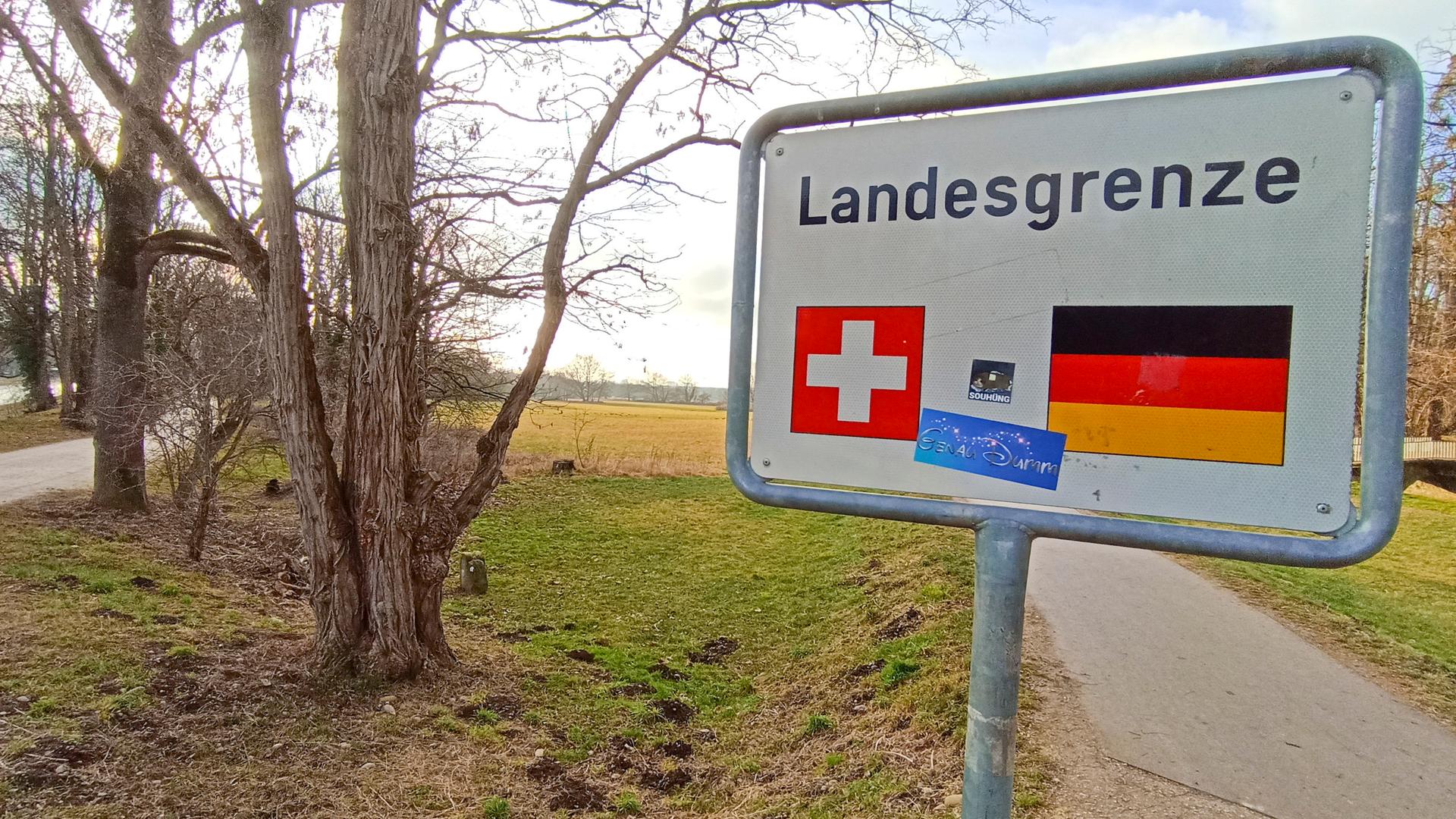 Das rechteckige weiße Schild mit dem Wort "Landesgrenze" und den Flaggensymbolen der Schweiz und Deutschlands steht an einem Feldweg vor kahlen Bäumen unter bewölktem Himmel.