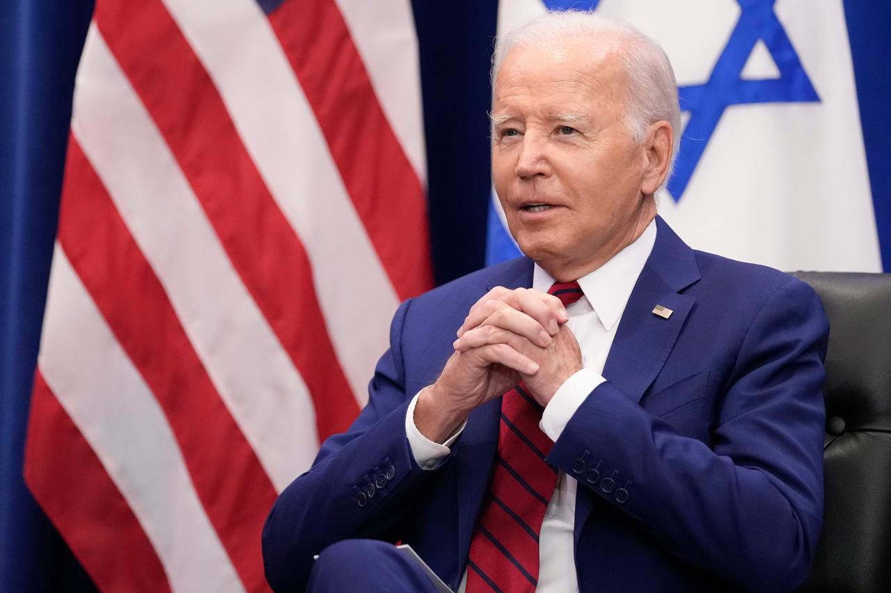 US-Präsident Biden sitzt und hat die Hände gefaltet, im Hintergrund zu sehen sind die Flaggen Israels und der USA.