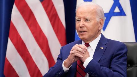 US-Präsident Biden sitzt und hat die Hände gefaltet, im Hintergrund zu sehen sind die Flaggen Israels und der USA.