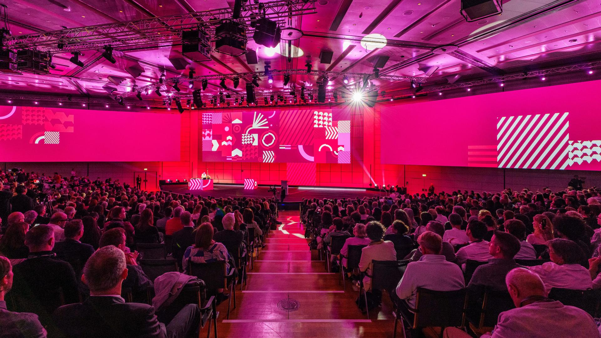 Das Bild zeigt eine Bühne und viele Sitzreihen. Der Raum ist in magentafarbenes Licht getaucht. Das Bild zeigt eine Bühne und viele Sitzreihen. Der Raum ist in magentafarbenes Licht getaucht.