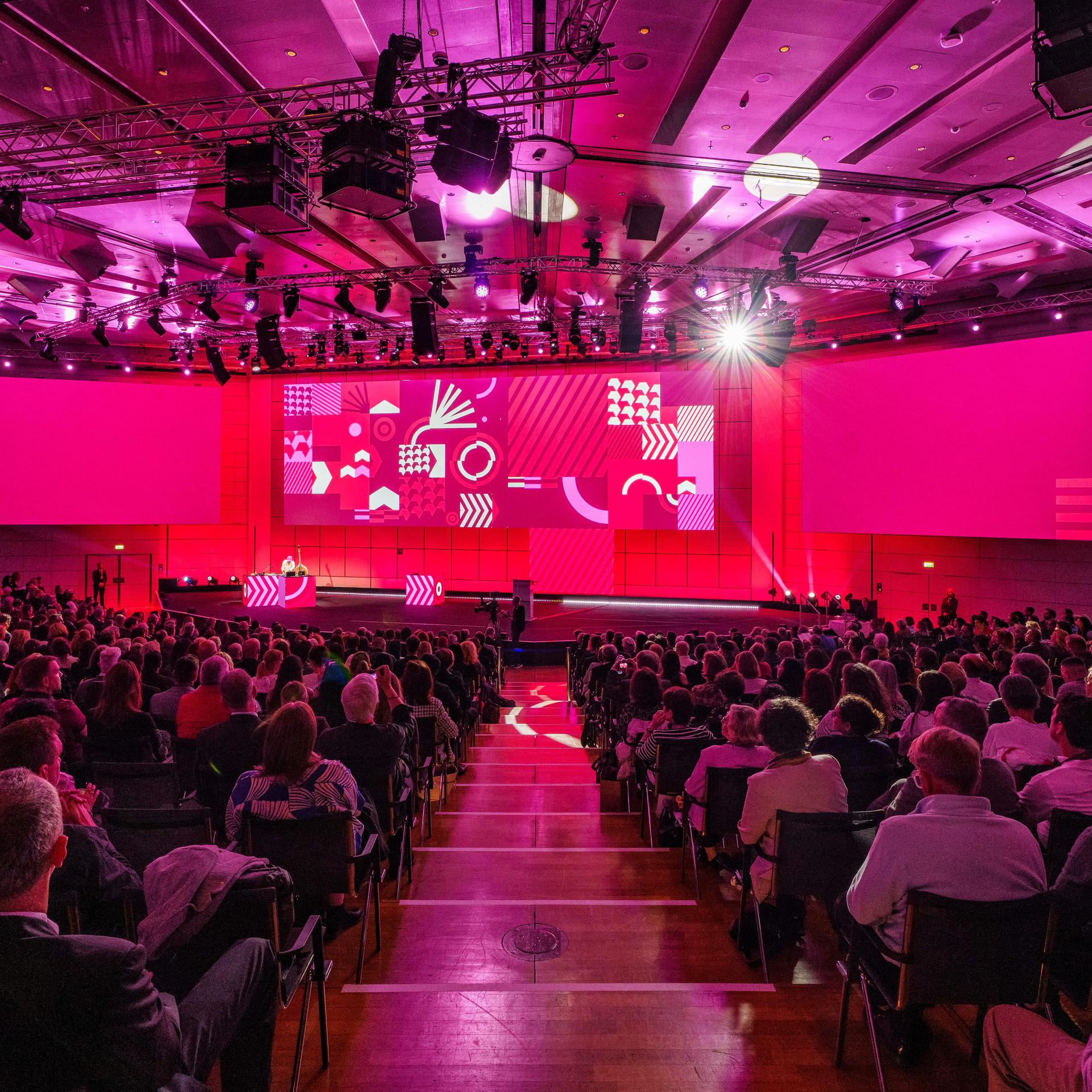 Das Bild zeigt eine Bühne und viele Sitzreihen. Der Raum ist in magentafarbenes Licht getaucht.