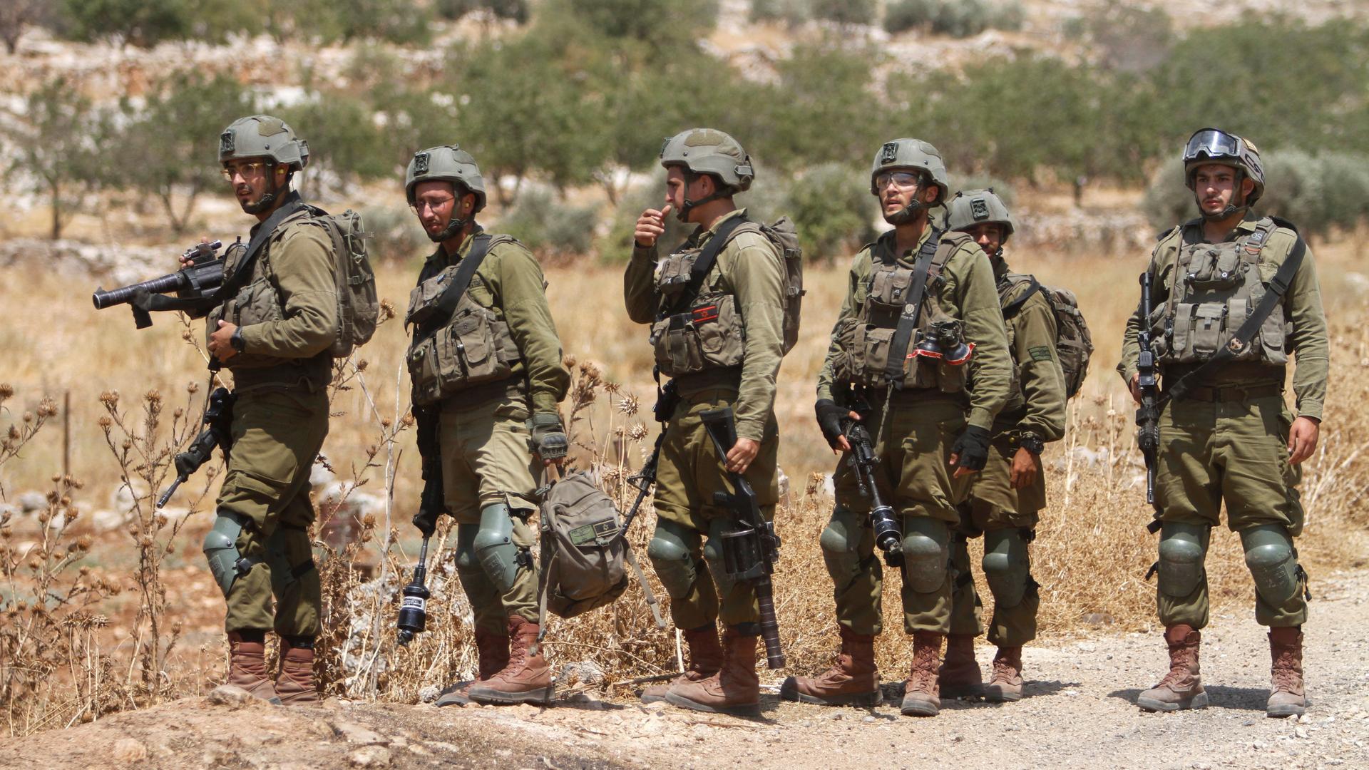 Die Soldaten mit Uniform, schussicheren Westen und Helmen stehen am Rande eines Feldweges, dahinter eine Fläche mit Olivenbäumen und verdorrtem Unkraut. Die Soldaten mit Uniform, schussicheren Westen und Helmen stehen am Rande eines Feldweges, dahinter eine Fläche mit Olivenbäumen und verdorrtem Unkraut.