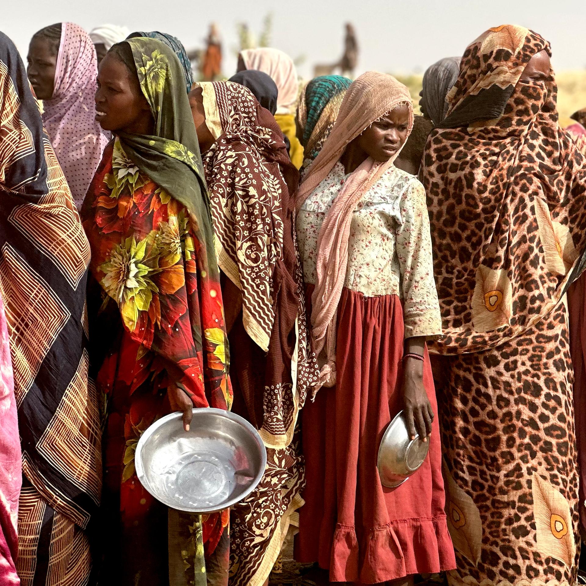 Frauen in bunten Gewändern stehen Schlange. Manche von ihnen halten metallene Schüsseln in den Händen.