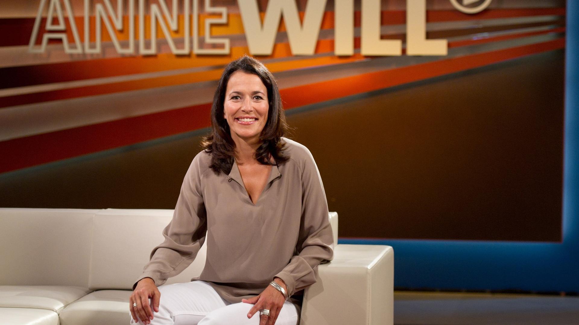 Die Journalistin Anne Will sitzt im Studio. Im Hintergrund ist der Schriftzug ihrer Talksendung "Anne Will" zu sehen. Sie trägt eine beigefarbene Bluse und eine weiße Hose.