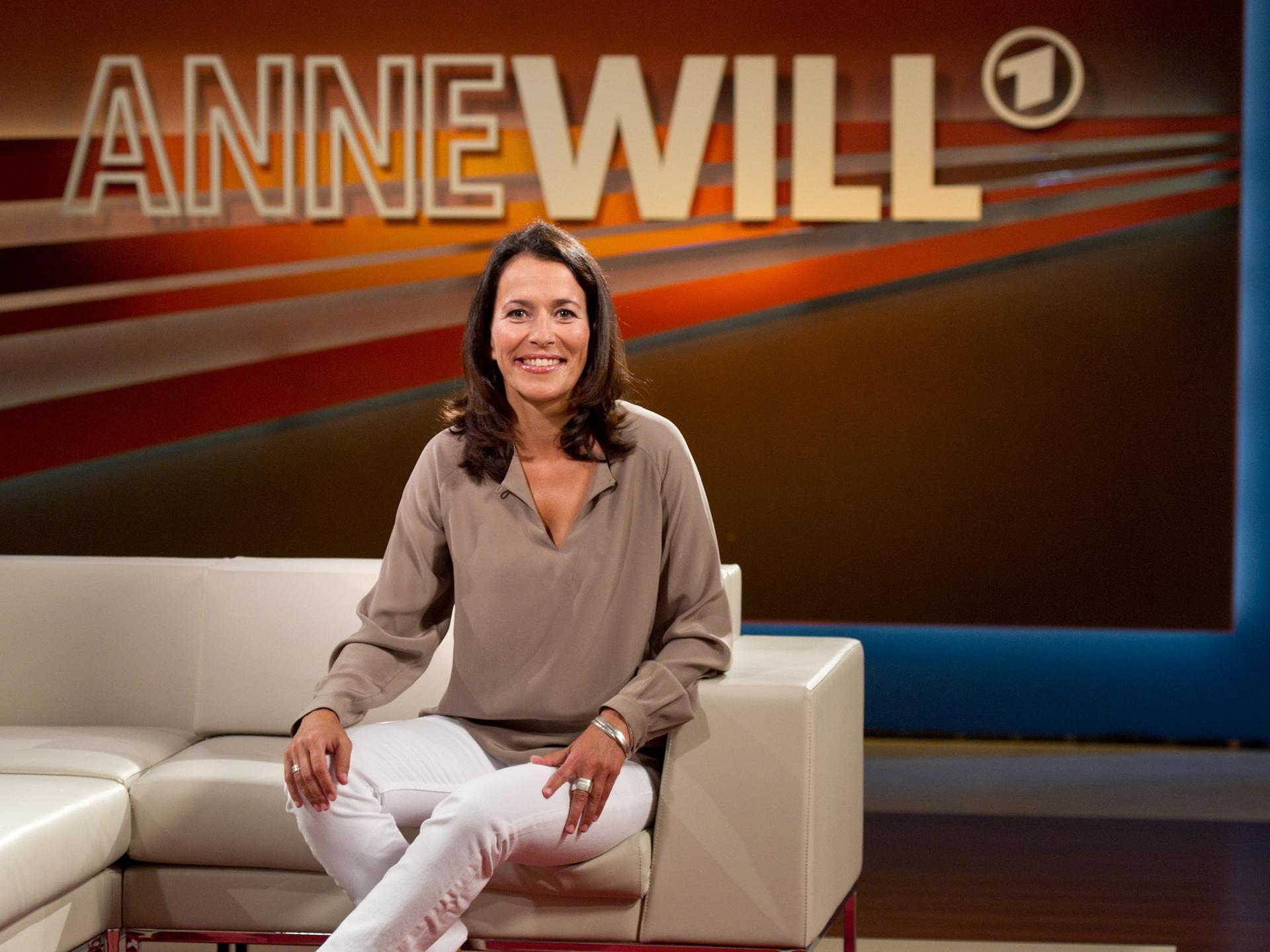Die Journalistin Anne Will sitzt im Studio. Im Hintergrund ist der Schriftzug ihrer Talksendung "Anne Will" zu sehen. Sie trägt eine beigefarbene Bluse und eine weiße Hose.