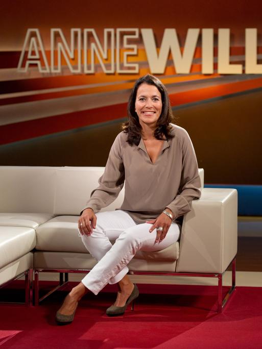 Die Journalistin Anne Will sitzt im Studio. Im Hintergrund ist der Schriftzug ihrer Talksendung "Anne Will" zu sehen. Sie trägt eine beigefarbene Bluse und eine weiße Hose.