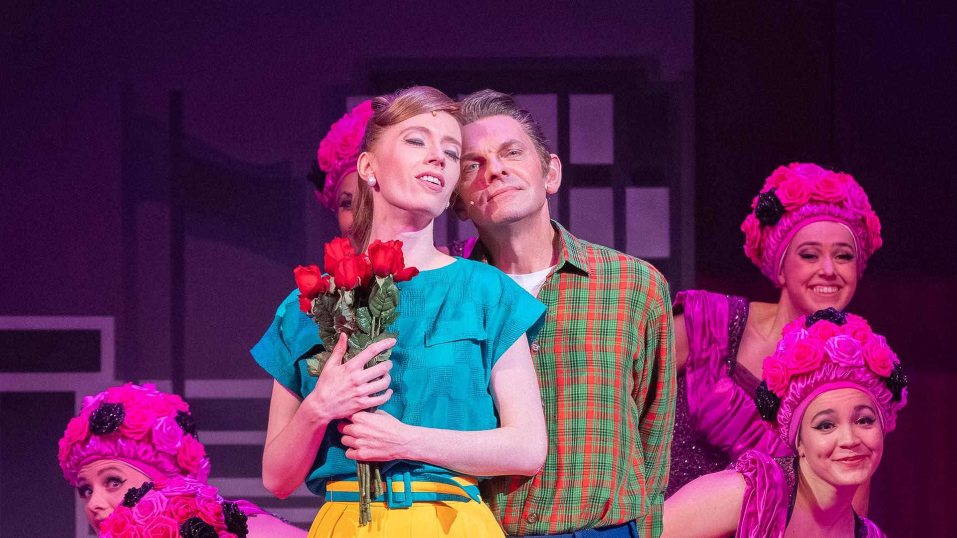 Eine junge Frau in gelbem Rock drückt Rosen an die Brust, während ein junger Mann hinter ihr steht. Im Hintergrund pinfarben kostümierte Tänzerinnen. Eine junge Frau in gelbem Rock drückt Rosen an die Brust, während ein junger Mann hinter ihr steht. Im Hintergrund pinfarben kostümierte Tänzerinnen.