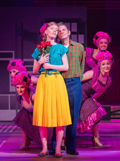 Eine junge Frau in gelbem Rock drückt Rosen an die Brust, während ein junger Mann hinter ihr steht. Im Hintergrund pinfarben kostümierte Tänzerinnen.