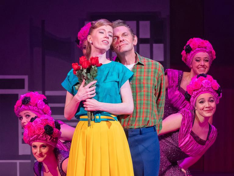 Eine junge Frau in gelbem Rock drückt Rosen an die Brust, während ein junger Mann hinter ihr steht. Im Hintergrund pinfarben kostümierte Tänzerinnen.
