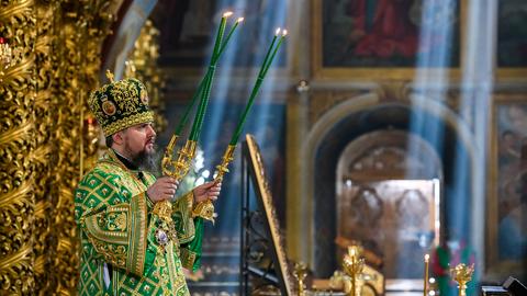Das Oberhaupt der orthodoxen Kirche der Ukraine hält zwei Kerzenständer mit grünen Kerzen in seinen Händen. Im Bildhintergrund sind einzelne Sonnenstrahlen zu sehen, die von oben in die Kathedrale einfallen.