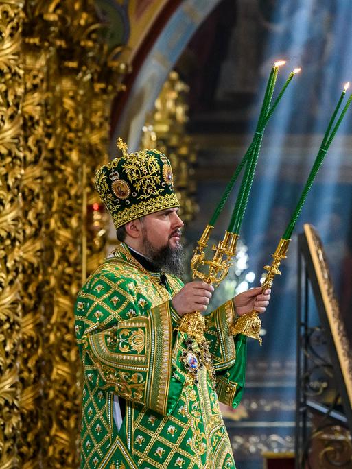 Das Oberhaupt der orthodoxen Kirche der Ukraine hält zwei Kerzenständer mit grünen Kerzen in seinen Händen. Im Bildhintergrund sind einzelne Sonnenstrahlen zu sehen, die von oben in die Kathedrale einfallen.