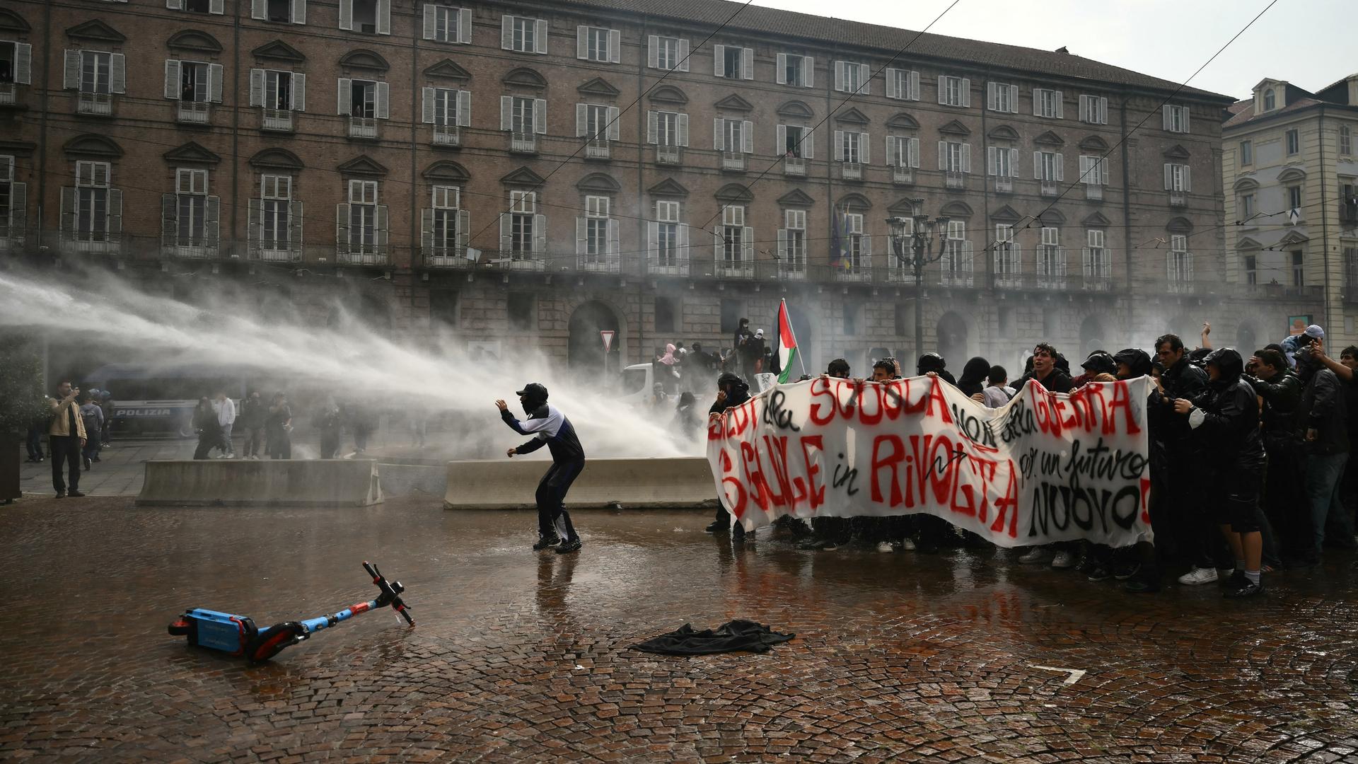 Die Polizei setzt bei einer Schülerdemo in Turin Wasserwerfer ein. Man sieht einen Wasserstrahl auf einem großen Platz. Rechts im Bild stehen durchnässte Schüler, die ein Transparent hochhalten. 