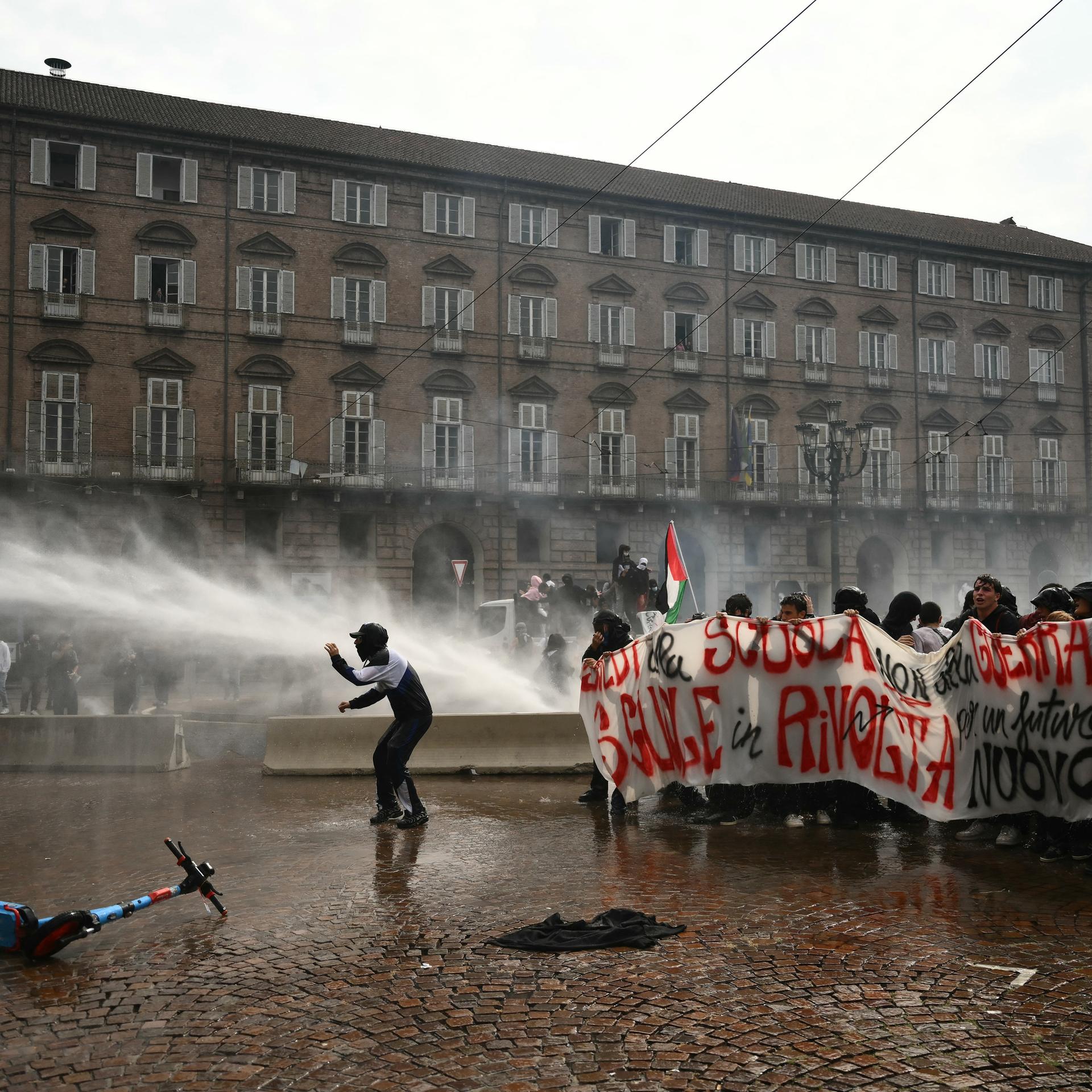 Die Polizei setzt in Turin Wasserwerfer ein. Man sieht einen Wasserstrahl auf einem großen Platz. Rechts im Bild stehen durchnässte Schüler, die ein Transparent hochhalten. 