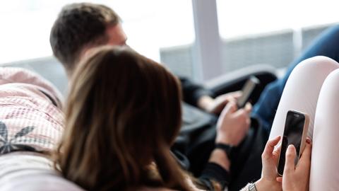 Eine junge Frau sitzt auf einem Sofa und benutzt ein Smartphone. Neben ihr sitzt ein junger Mann, der ebenfalls am Handy tippt. 
