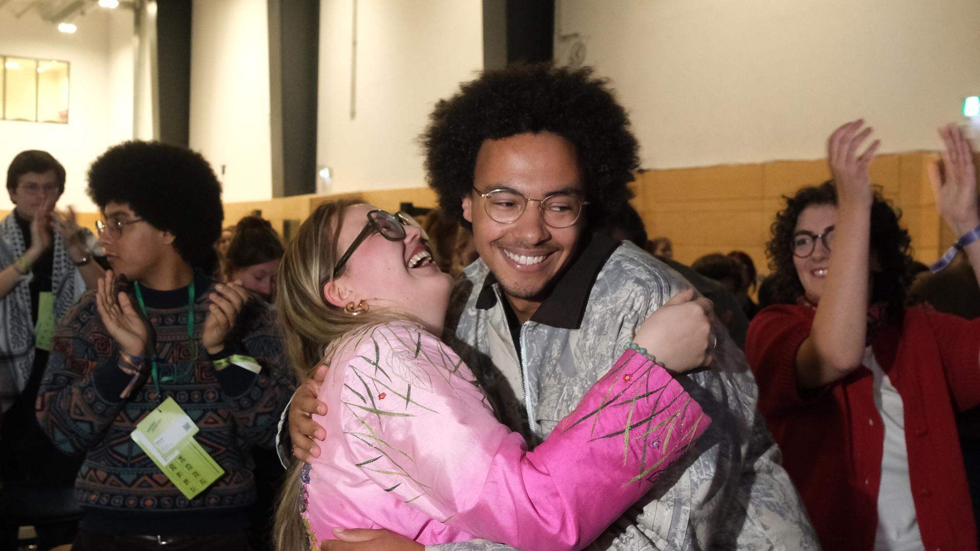 Das Bild zeigt Luis Bobga und Henriette Held kurz nach ihrer Wiederwahl. Beide umwarmen sich und lachen. Im Hintergrund sind jubelnde Parteimitglieder zu sehen.