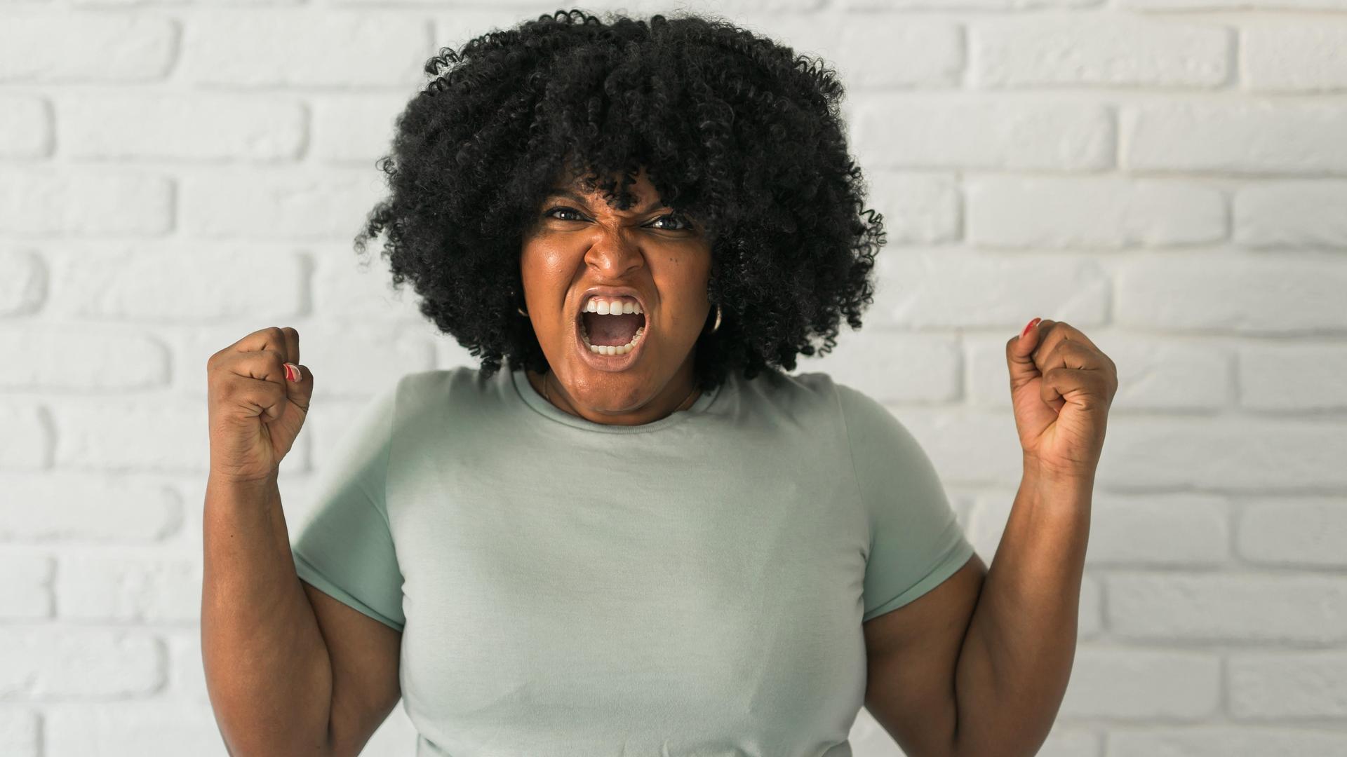 Eine Frau trägt eine Afro-Frisur und einem grauen T-Shirt ballt die Fäuste und schreit wütend mit in Richtung der Kamera