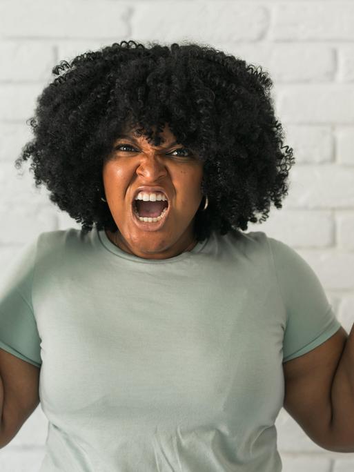 Eine Frau trägt eine Afro-Frisur und einem grauen T-Shirt ballt die Fäuste und schreit wütend mit in Richtung der Kamera