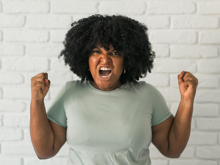 Eine Frau trägt eine Afro-Frisur und einem grauen T-Shirt ballt die Fäuste und schreit wütend mit in Richtung der Kamera