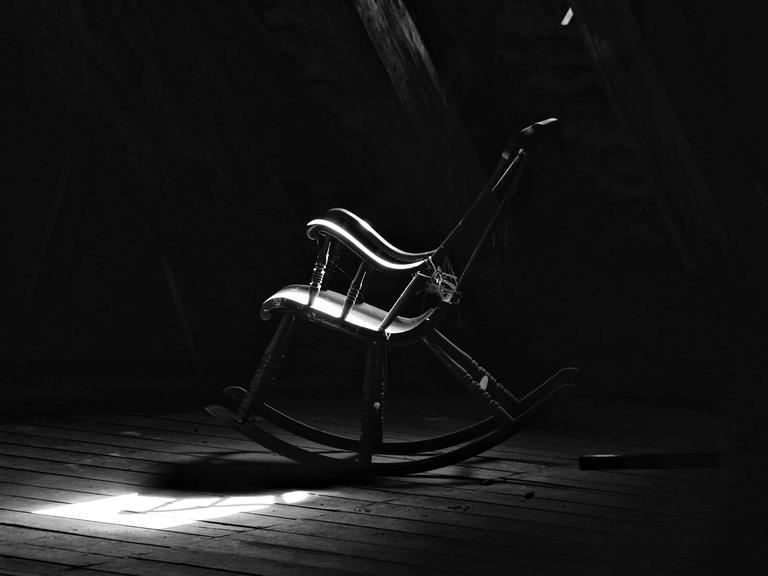 Schwarz-Weiß-Bild eines verstaubten Schaukelstuhls auf einem Dachboden.