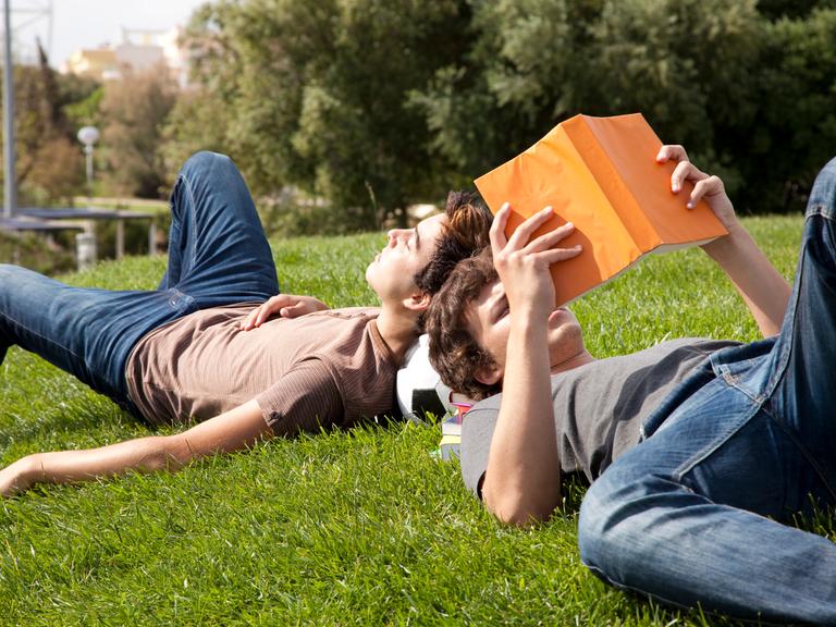 Zwei junge Personen liegen auf einer Wiese, neben ihnen Bücher und ein Fußball Zwei junge Personen liegen auf einer Wiese, neben ihnen Bücher und ein Fußball