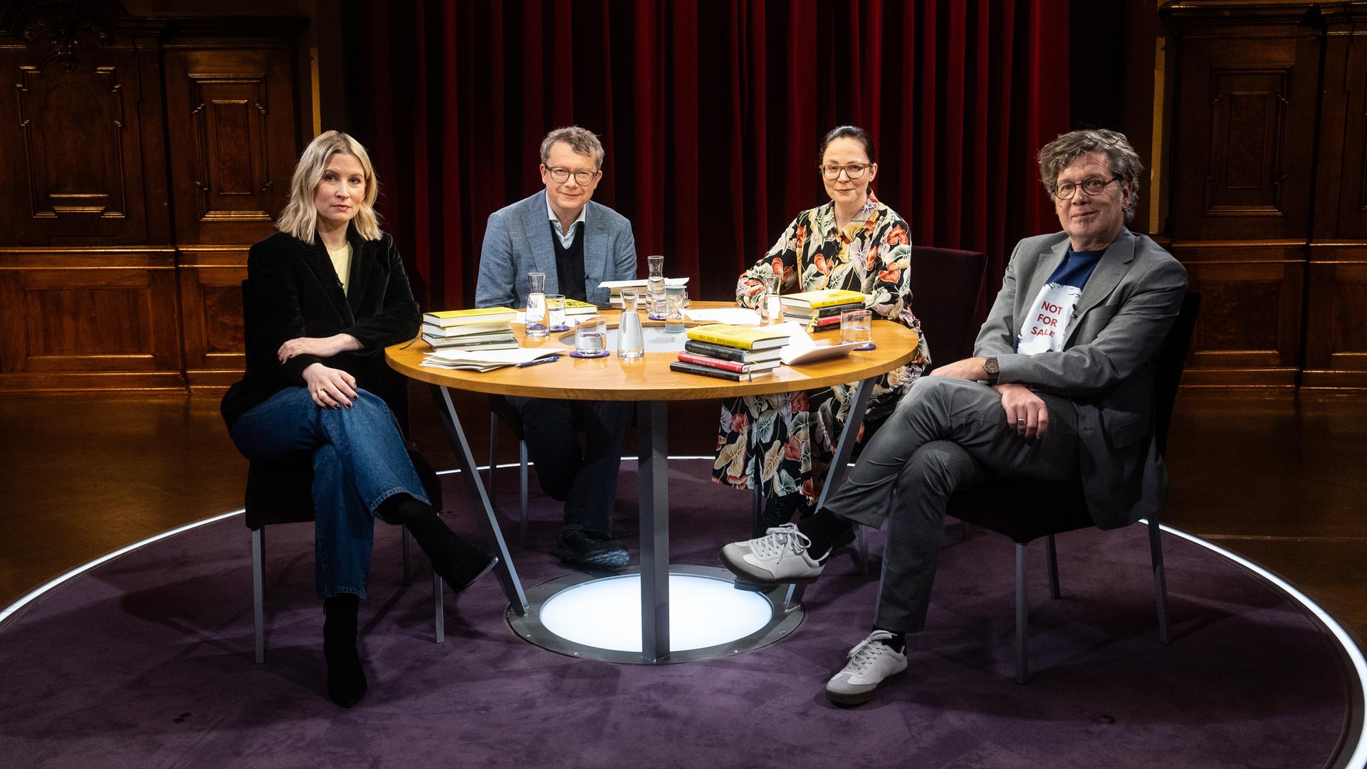 Mara Delius, Adam Soboczynski, Thea Dorn und Franzobel sitzen an einem runden Tisch, auf dem die Bücher der Sendung liegen, und schauen in die Kamera.