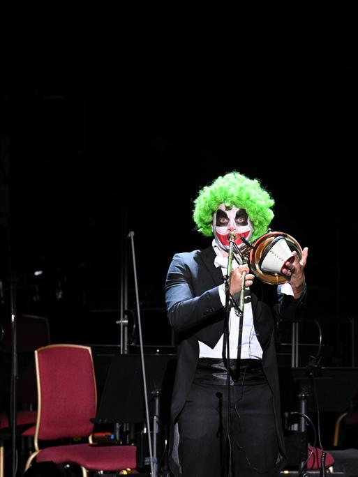 Ein als Clown geschminkter Posaunist hält den Dämpfer an sein Instrument und spielt