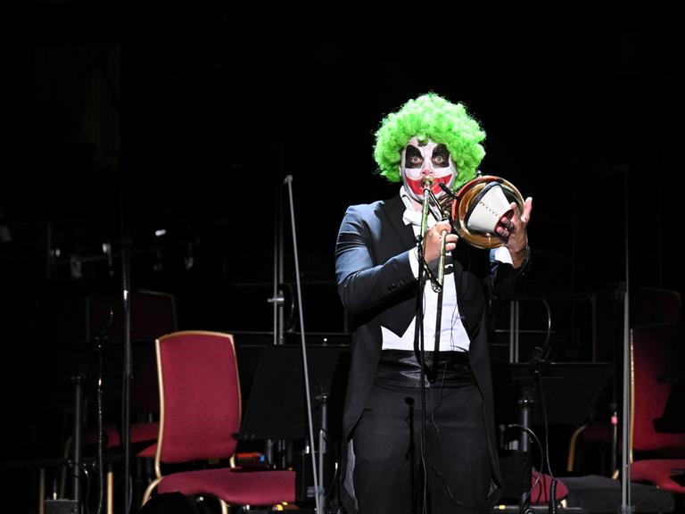 Ein als Clown geschminkter Posaunist hält den Dämpfer an sein Instrument und spielt