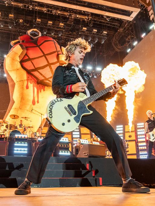 Billie Joe Armstrong von Green Day steht auf der Bühne. In der Hand hält er eine Guitarre. Im Hintergrund sind Feuer zu sehen. 