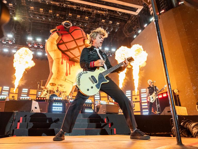 Billie Joe Armstrong von Green Day steht auf der Bühne. In der Hand hält er eine Guitarre. Im Hintergrund sind Feuer zu sehen. 