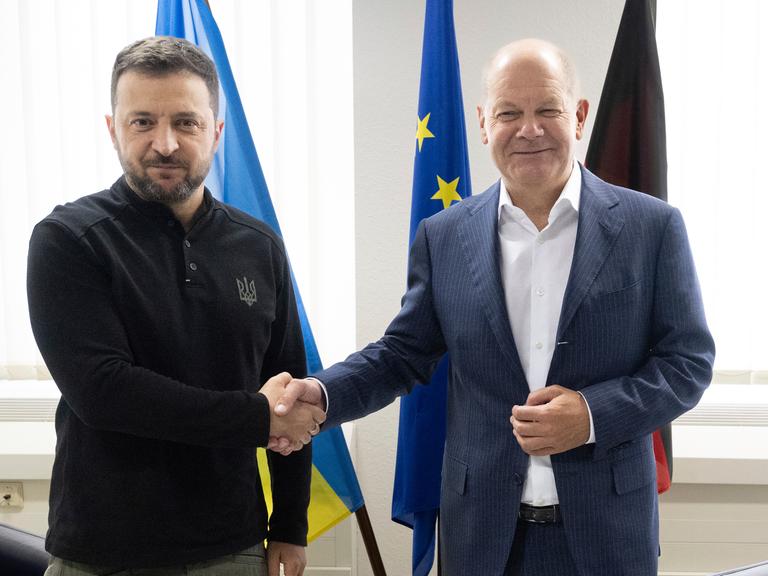 Bundeskanzler Olaf Scholz (SPD, rechts), und der ukrainische Präsident Wolodymyr Selenskyj geben sich bei einem Treffen die Hände und schauen in die Kamera.