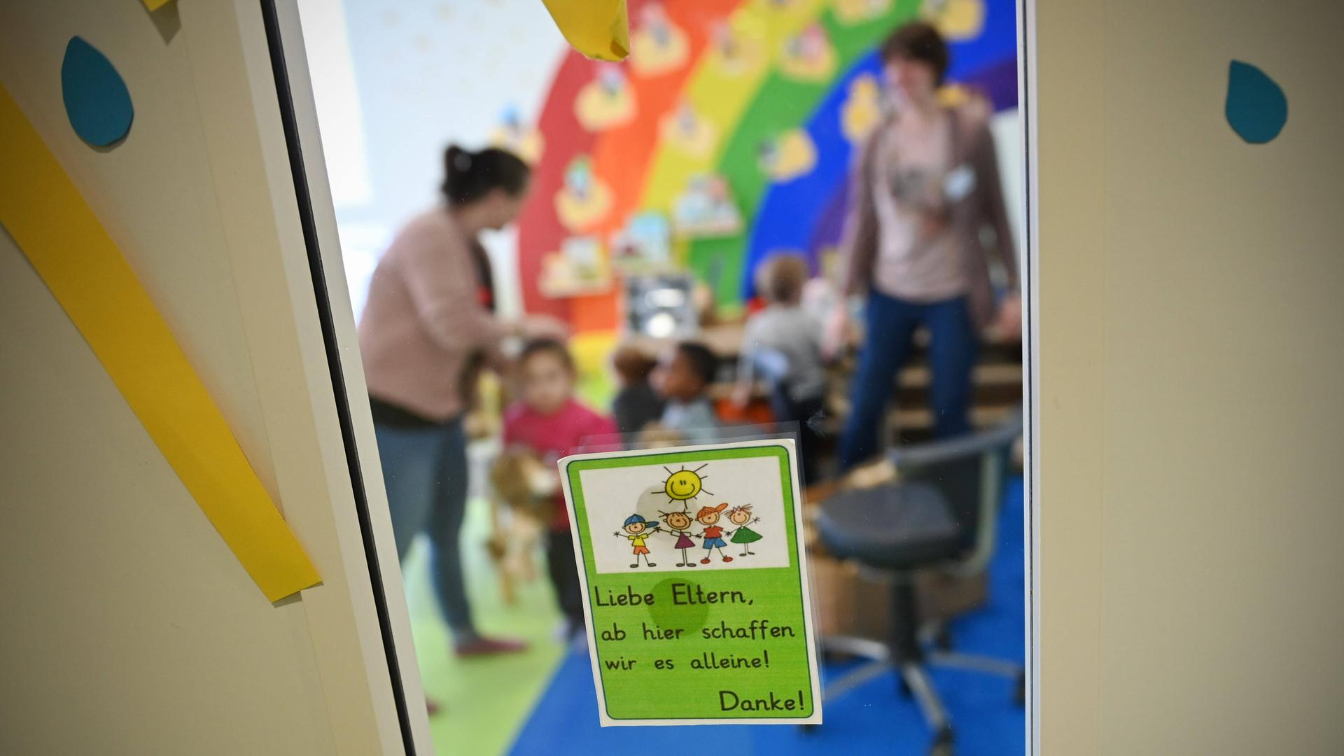 Kinder mit Erzieherinnen in einer Kita. Scharf im Vordergrund ist ein Hinweiszettel an der Glastür zu sehen. Die kinder und Erzieherinnen in der Kita sind verschwommen und nicht zu erkennen.