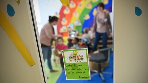 Kinder mit Erzieherinnen in einer Kita. Scharf im Vordergrund ist ein Hinweiszettel an der Glastür zu sehen. Die kinder und Erzieherinnen in der Kita sind verschwommen und nicht zu erkennen.