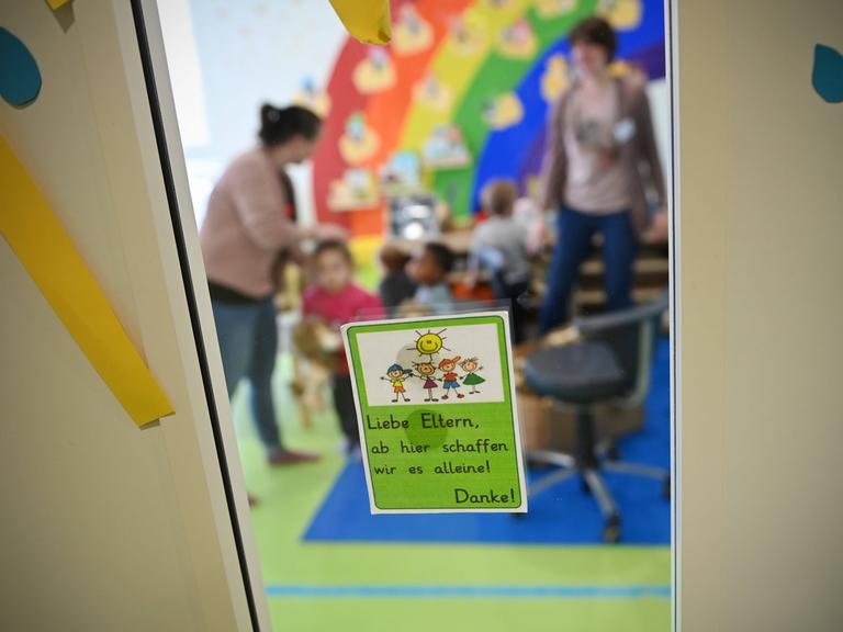 Kinder mit Erzieherinnen in einer Kita. Scharf im Vordergrund ist ein Hinweiszettel an der Glastür zu sehen. Die kinder und Erzieherinnen in der Kita sind verschwommen und nicht zu erkennen.