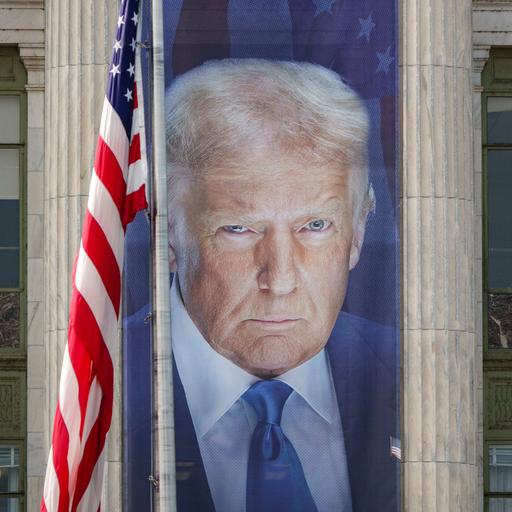 Ein Banner mit einem Bild von US-Präsident Donald Trump hängt an der Fassade des US-Landwirtschaftsministeriums zwischen zwei Säulen. Daneben hängt die US-amerikanische Flagge. 