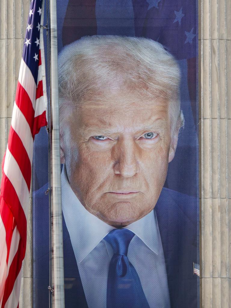 Ein Banner mit einem Bild von US-Präsident Donald Trump hängt an der Fassade des US-Landwirtschaftsministeriums zwischen zwei Säulen. Daneben hängt die US-amerikanische Flagge. 
