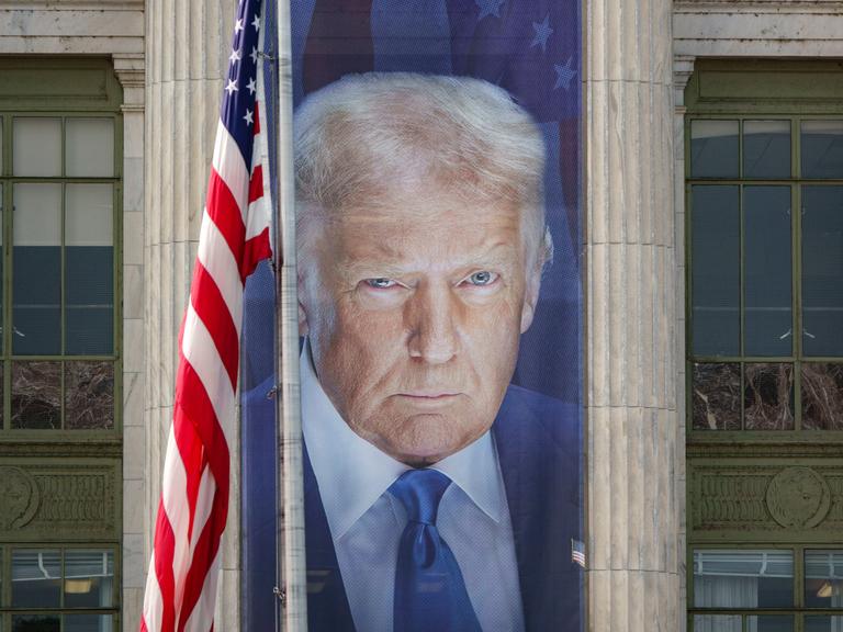 Ein Banner mit einem Bild von US-Präsident Donald Trump hängt an der Fassade des US-Landwirtschaftsministeriums zwischen zwei Säulen. Daneben hängt die US-amerikanische Flagge. 