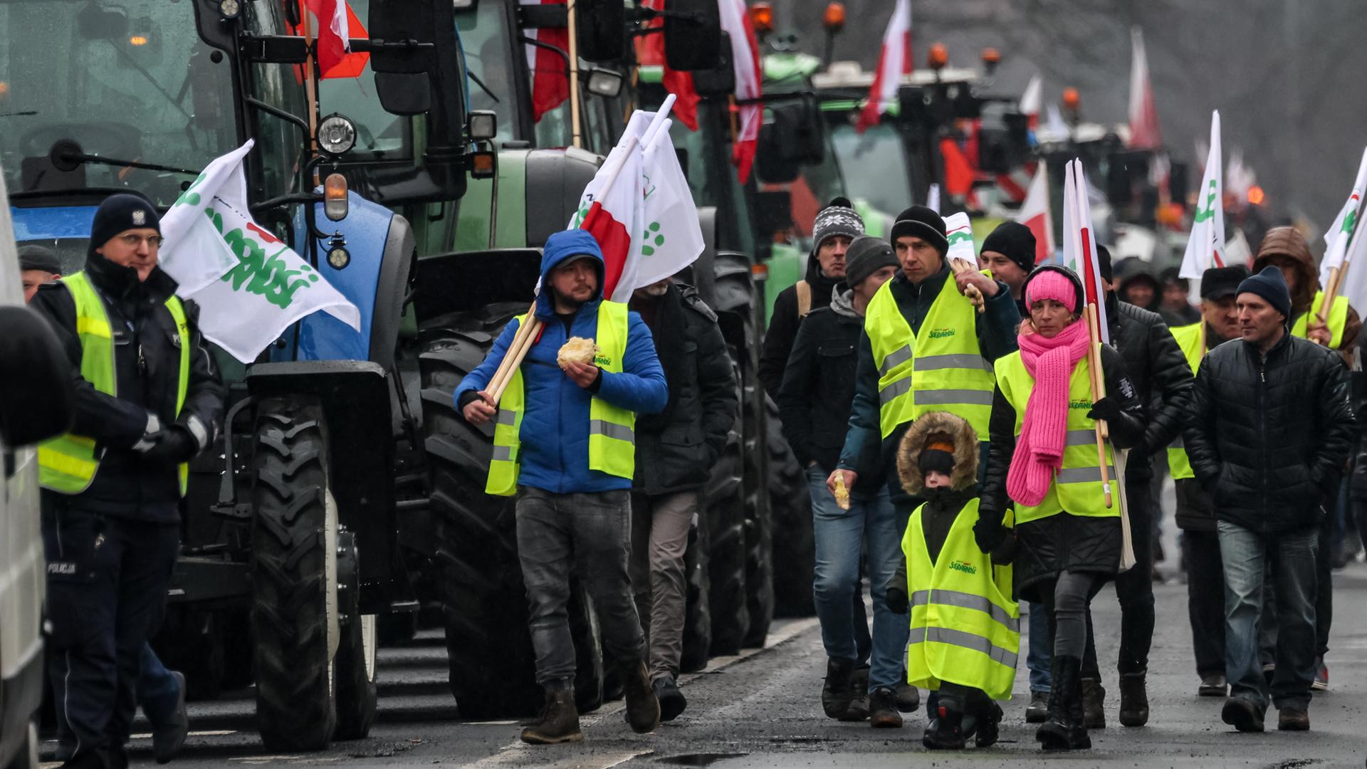 Bauern in gelben Warnwesten und mit Protestflaggen laufen an geparkten Traktoren vorbei.