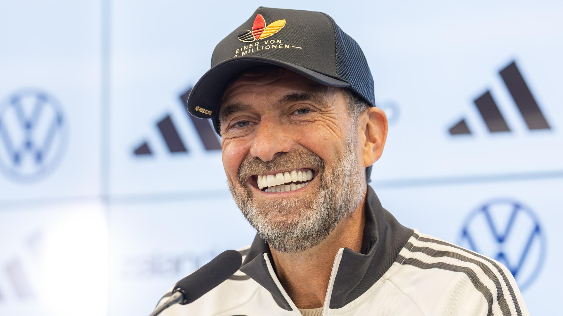 Bundestrainer Jürgen Klopp sitzt vor einem Mikrofon und lacht. Er hat eine Trainingsjacke an und ein Käppi auf dem Kopf. Bundestrainer Jürgen Klopp sitzt vor einem Mikrofon und lacht. Er hat eine Trainingsjacke an und ein Käppi auf dem Kopf.