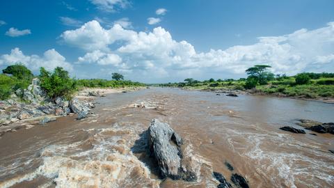 Der Mara Fluss in der Nähe der Grenze zwischen Kenia und Tansania, Im Vordergrund fliesst Wasser über Steine, im Hintergrund und seitlich ist Landschaft der Serengeti zu erkennen.
