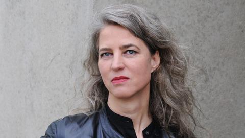 Eine Frau mit lockigem, grauem Haar und blauen Augen steht vor einer grauen Wand. Sie trägt eine schwarze Jacke in Lederoptik.