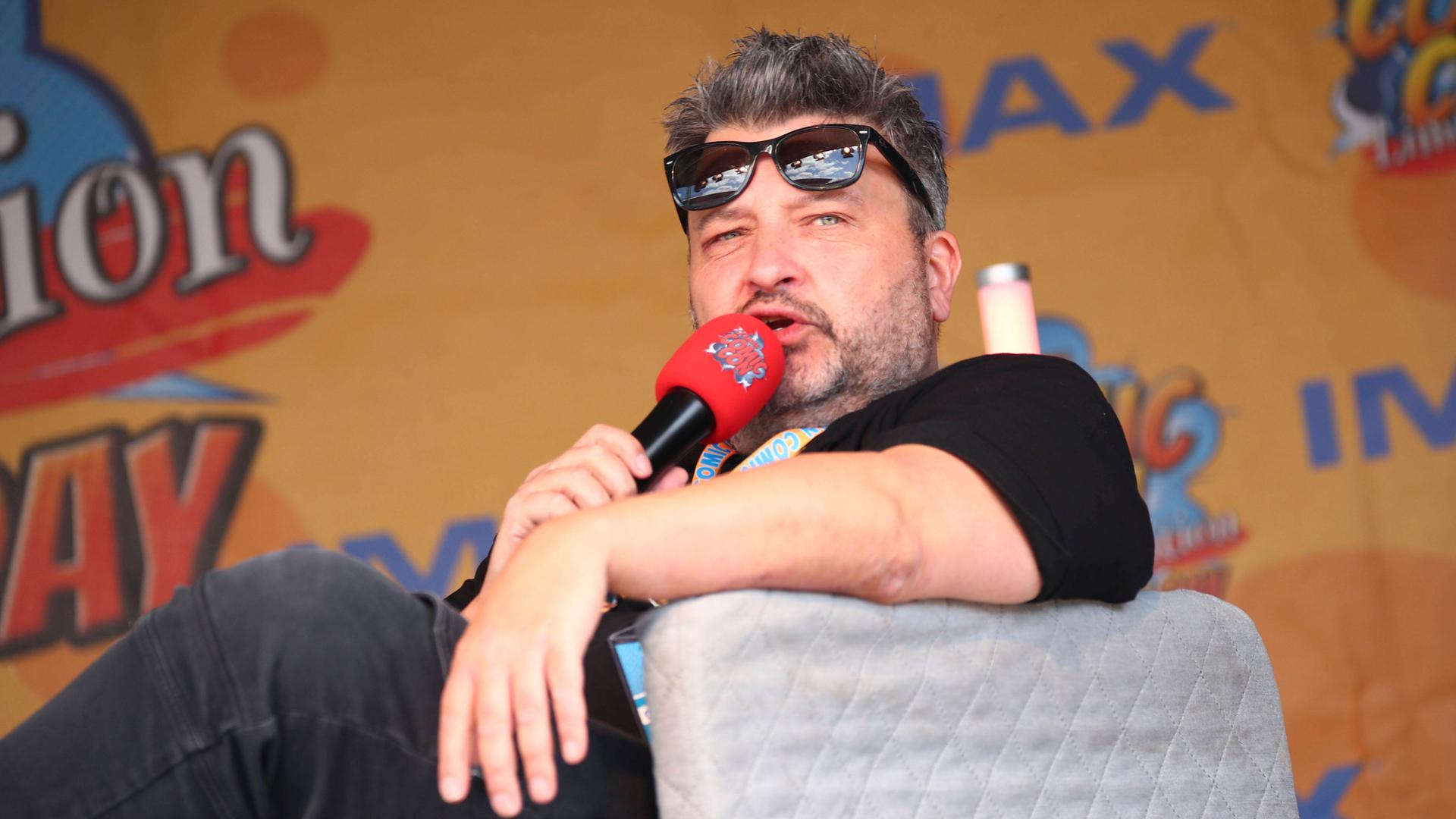 Der Comiczeichner Oli Hilbring hat die Sonnenbrille auf der Stirn und spricht in ein Mikrofon während er in einem Sessel sitzt.