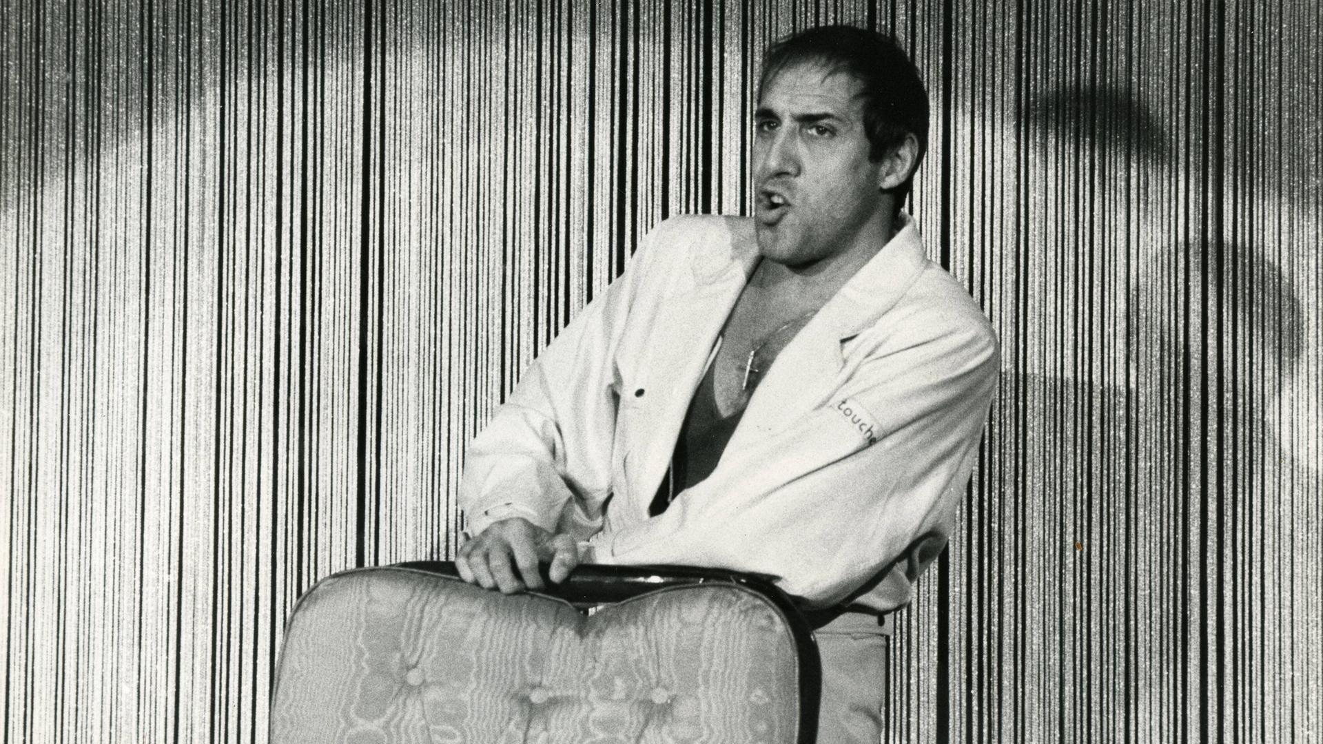 Schwarzweißaufnahme von Adriano Celentano, der lässig an einen Sessel lehnt und singt.