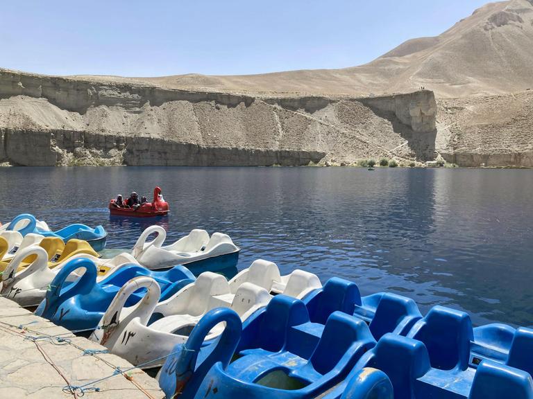 Ein See in einer Wüstenregion. Viele Tretboote liegen an seinem Rand. Eines fährt darauf herum. Die Tretboote haben das Aussehn von Schwänen. Sie sind blau, weiß und auch ein gelbes ist zu sehen.