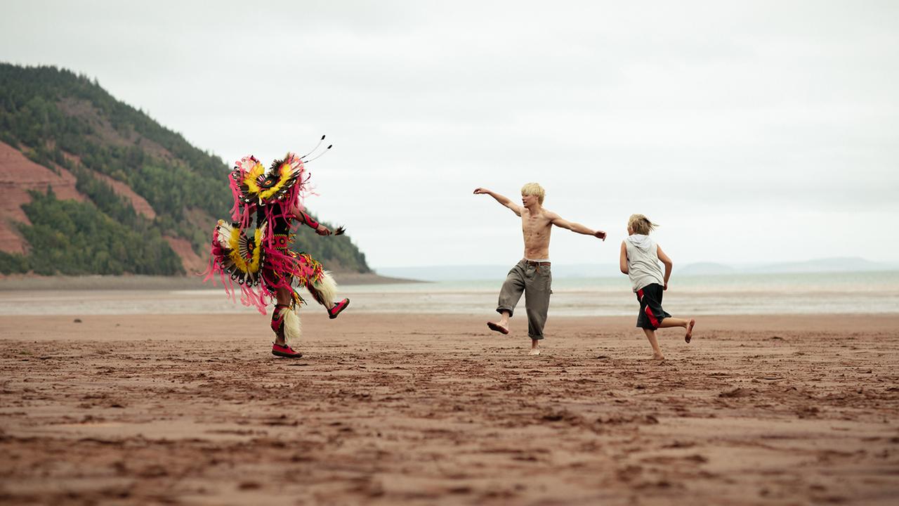 Eine Filmszene aus "Wildhood" zeigt drei Personen an einem sonst menschenleeren Meeresstrand. Zu sehen sind ein Mann in indigener Kostümierung und zwei Jungen beim Tanzen.